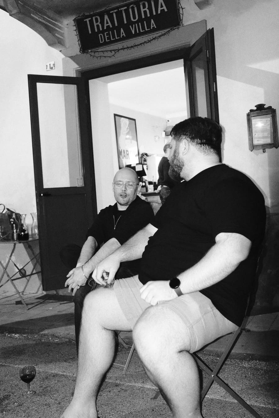 Two teammates chatting outside a trattoria, one holding a glass of red wine