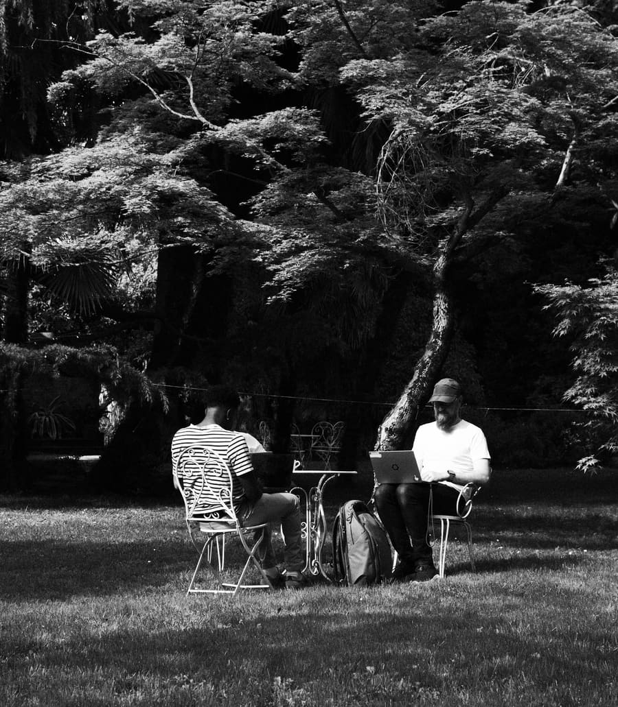 Two teammates working outdoors under a tree, one on a laptop with a backpack nearby