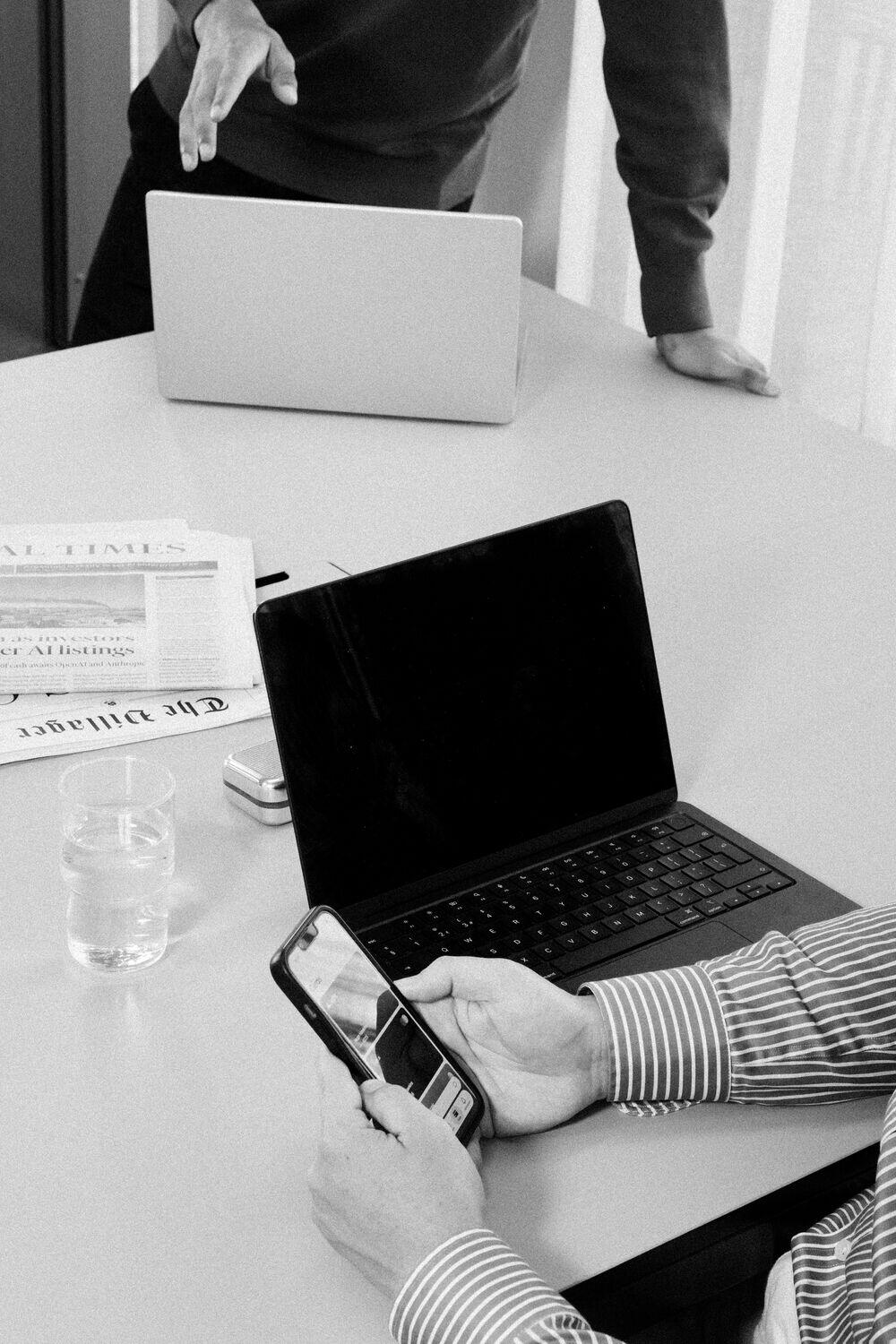 A desk with a newspaper, a laptop, and a phone in hand